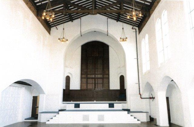 After: Interior view of church sanctuary and altar. All wall surfaces are painted white. The original wood floor has been refinished with a dark stain. Beamed ceiling is visible.